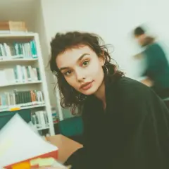 Młoda kobieta w bibliotece, otoczona książkami. Czyta, a obok niej widać ruchliwą postać. Ibiden, co to za miejsce?