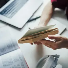 Ręce trzymają otwartą książkę, obok laptop i słuchawki. To dobry przykład, jak tworzyć bibliografię.