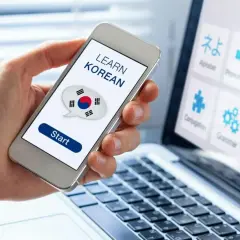 Dłoń trzyma smartfon z napisem "LEARN KOREAN" i flagą Korei. Obok laptop z ikonami do nauki języka koreańskiego od podstaw.